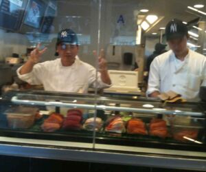 Sushi chefs prepare California Rolls and flash the peace sign behind glass windows.